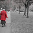The back view of an old lady wearing a red overcoat & black skirt, walking along a grey mono-toned suburban tree-lined path.