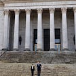 Photo of the sandstone Capitol building, with Judge Yu and the governor talking together while walking down a wide, tall set of steps with the soaring sandstone columns of the building behind them