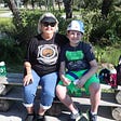 Two family campers, Dawn and Caden, smile while Caden waits to climb the rock wall at Camp Aranzazu. Dawn and Caden are seated at log benches in the middle of camp