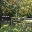 Near the left, a wooden sign reads “Unity Park” in large white letters, followed by “M-NCPPC Montgomery County” and the street address of “3724” below. The sign is on a grassy area. To the right is a wooden fence with a road just visible. Behind the sign, a wooded area is visible.