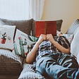 A man reclining on a couch with pillows that have cactuses on them. He has a red book opened and covering his face.