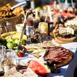 A large spread of food on a table covered in white cloth. Cheeses, crackers, cured meats, and fruit.