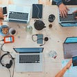 Multiple laptops, phones, food, and glasses on a table with three people working.