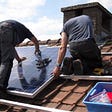 Color photo of two men installing solar panels on a roof.