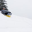 Young person on yellow sled happily heading downhill with tree in background