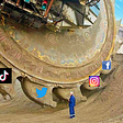 A giant digging machine with social media company logos added to the buckets extracting huge amounts of dirt from the earth with a man standing in front of it totally dwarfed by the machine’s size.