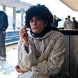 Curly haired woman in a sweater sitting at a train station smoking a cigarette and staring into the camera