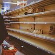 Photo of almost empty shelves due to Covid in a Swiss grocery store. A person is walking on the left with a red basket filled with items.