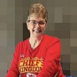 A smiling woman with brown hair with grey in it, wearing glasses and a red Kansas City Chiefs long sleeved shirt.