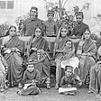 Image description: A group of Bene Israel Jewish community. Children are seated on the floor in the front, women are seated on chairs, and men and older boy are standing behind women. The women are wearing saris and traditional Indian jewelry like nose rings, earrings, necklaces, and anklets, and are holding babies. The children and men are wearing traditional Jewish hats with flat tops that cover the crown of their heads.