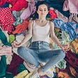 Young girl laying amidst a bed of tossed clothing in a supine lotus position, meditating with a smile on her face.