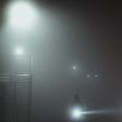 A man carrying a flashlight explores a foggy night. Two lights hang in the air a short distane behind him. They almost look like eyes. A metal staircase is to the left of the image. Maybe they should not stray so far into the unknown.