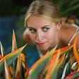 Woman with blond hair, eyes looking to the right, playfully crouched behind orange and green plants.