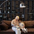 Mother in a hijab and daughter reading on a couch in front of bookshelves.