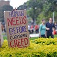 A banner reads “Human needs before corporate greed”