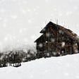 Photo of an old wooden cabin on a snowy hilltop beneath a grey cloudy sky.