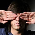 Man covering eyes with hands. One hand has a dollar sign and the other says, lie.