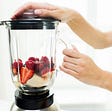Photo of woman’s hands on blender with fruit and cream inside.