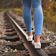 person wearing blue jeans and white tennis shoes walking on edge of railroad track