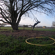 A girl stretches using two gym rings hanging off a tree silhouetted against late afternoon sky. A yellow hula hoop lies on the grass.