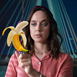Image from the Netflix show Sexify. A white blue eyed brunette stares at a vibrator, a look of challenge in her eyes. The vibrator image is covered by a banana.
