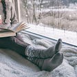A woman relaxing and reading by an open window with a serene snowscape outside.