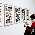 A woman in an art gallery looking at a display of various 2D matrix barcodes.