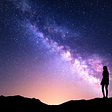 A photo of a silhouette of standing woman looking at the Milky Way Galaxy at night. The woman is standing on a desert landscape. A multitude of stars light up the night sky.