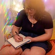 Photo of Allison writing in her journal. She is wearing a dark colored outfit and crystal necklace while sitting in a cozy nook of pillows. There is a photography effect used that gives the photo a blurred quality in spots and casts lines of rainbow prisms in others.