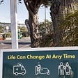 A sign reading, “Life can change at any time”, empty street and tree in background