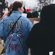 Image of the back of feminist marchers. One marker is wearing a blue denim jacket with a DIY-looking slogan on its back saying “GRL PWR” and a face with abstract tears coming from it in straight lines. The other marchers are in dark colors.