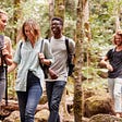 Four people with backpacks share a laugh while walking through a wooded area.