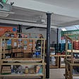 Transition Stirling has a range of reuse and recycle projects including the distribution of unused food from supermarkets and a tool library. Pictured is a large room with white walls, a black pillar in the centre and a series of hand made wooden market stalls with bunting and banners in bright colours