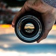 Image of a camera lens up against a blurry landscape, with the inner eye of the lens reflecting a clear landscape.