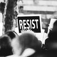 Black and white photograph of a protest, the only thing in focus is a sign that says RESIST.