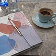Colourful notebook with a pen and a blue cup of espresso on a marble countertop