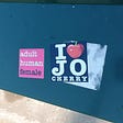 Two colourful stickers on a metal surface outdoors. One reads “Adult Human Female”, while the other reads “I [heart] Jo Cherry”