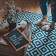 Photo of geometric patterned rug and crate coffee table with a placard on it reading “Sorry for the things I’ve said when it was winter.” The legs of a white woman wearing distressed jeans and white slippers are off to the side..