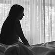 A woman sits in shadow on a bed. She is alone in a white room.