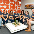 Seven members of the Architectural Designs team pose in an office with a bright orange and write wall. They are wearing blue shirts that read Bueno to honor AD alumnus Ryan Bard, who is pursuing his lifelong dream of becoming a commercial pilot.