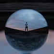 A glass ball whose reflective surface shows a single man standing in the middle of an empty street.
