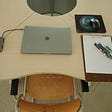 A chair and table with  writing pad and paper, pens, pencil and eraser, computer and mouse.