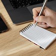 Cropped person writing in a small notepad with a pen on their workspace