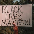 Black person’s hand shown holding sign that reads “Black Lives Matter.”