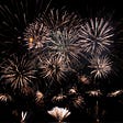 A cluster of six gold brocade effectfireworks surrounded by smaller firework explosions on a black sky background