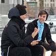 Two young men conversing while sitting on a park bench on winter day