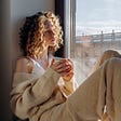 A woman with honey colored medium length curly hair sitting, looking out a window. She is holding a mug wearing a beige sweater, pants, and white tank top.