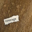 A close view of a wooden table or floor. A scrap of torn off-white paper with the word COURAGE typed on it.