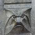 A frightening stern face, a mascaron, carved in gun-metal gray stone peers out from a doorway in Amsterdam. Photo by author.
