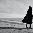 Greyscale image. Person in a black cloak stands alone on a beach near the edge of the water looking out, their shadow extending behind them.